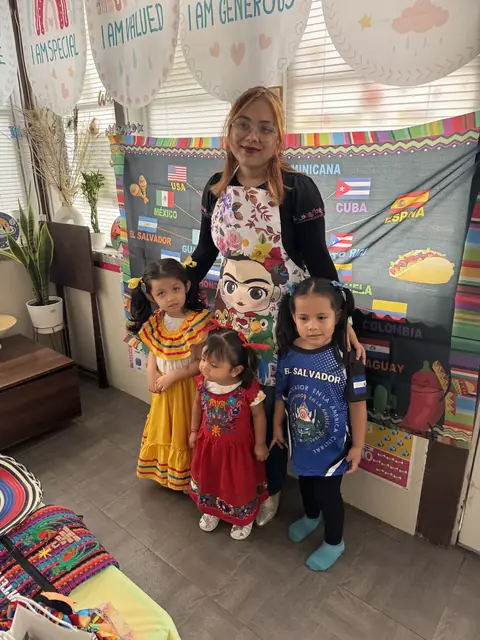 A caregiver in a floral apron stands with three girls in traditional dresses and an El Salvador jersey