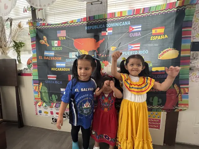 Three girls in traditional dresses and an El Salvador jersey dance in front of a map of Latin America