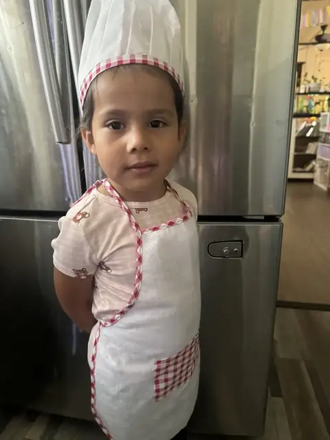 A toddler in a chef hat and gingham apron stands in the kitchen