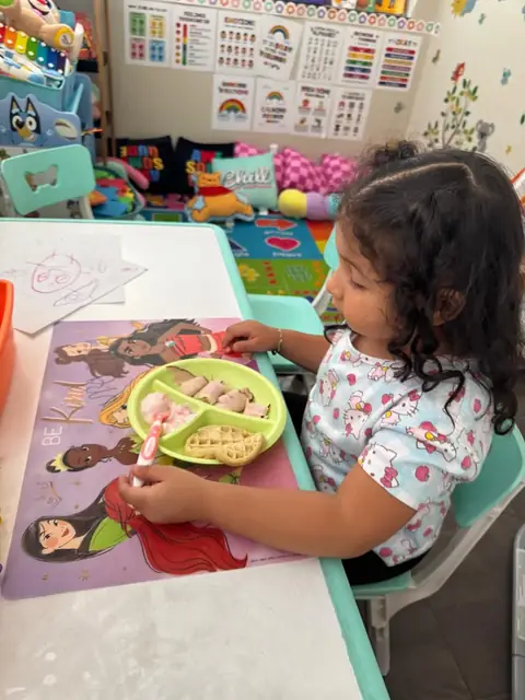 A toddler eats a lunch of fruit, rolled turkey and a waffle at a small table