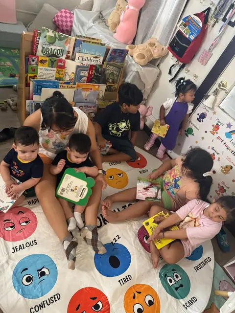 A caregiver reads with two babies on her lap while older children look at books
